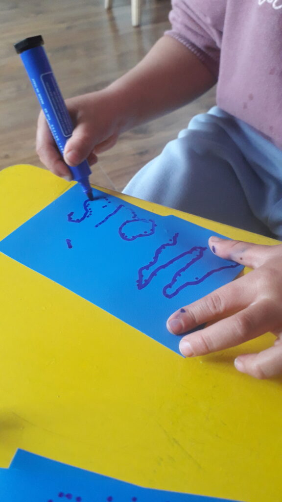 Ein Kind schreibt ''Mais'' auf ein blaues Papier