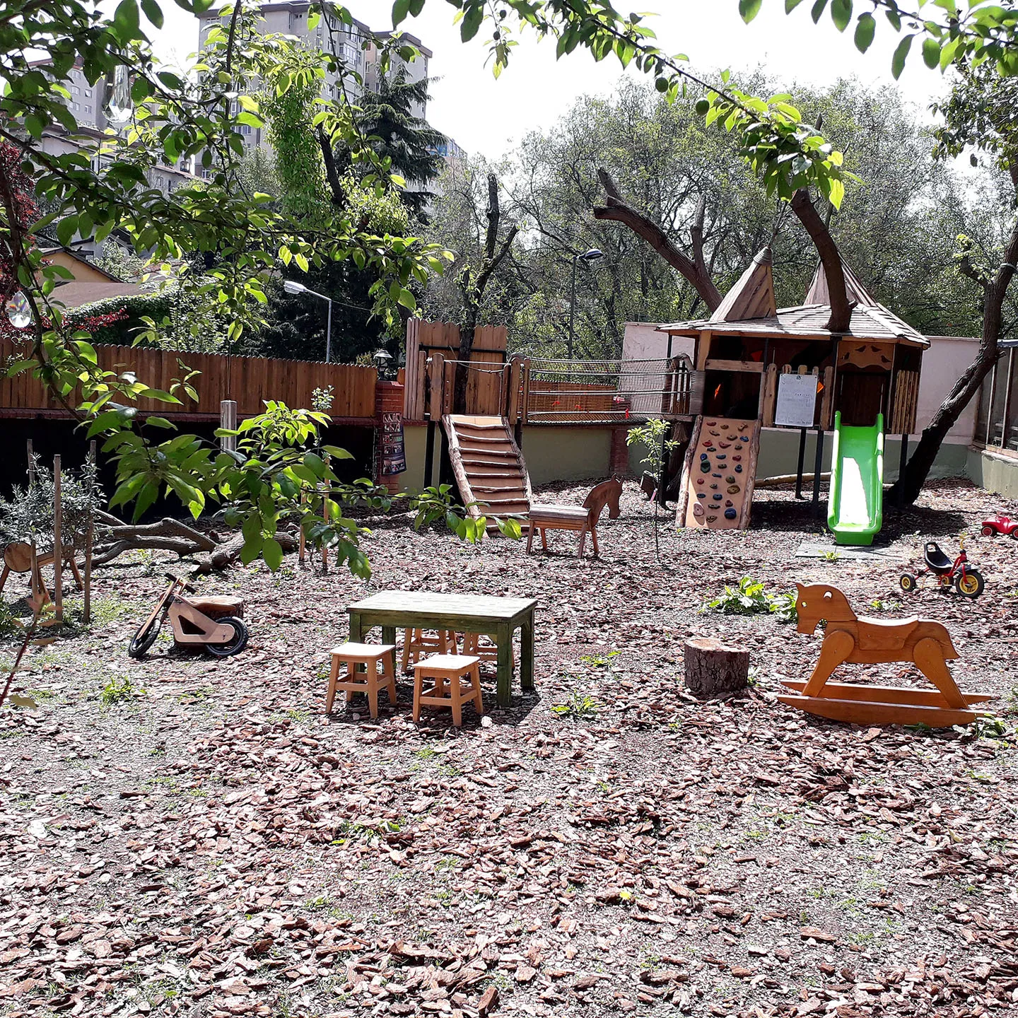Der Blick auf den EBİ Garten mit Tischen, Hocker, Schaukelpferd und Baumhaus