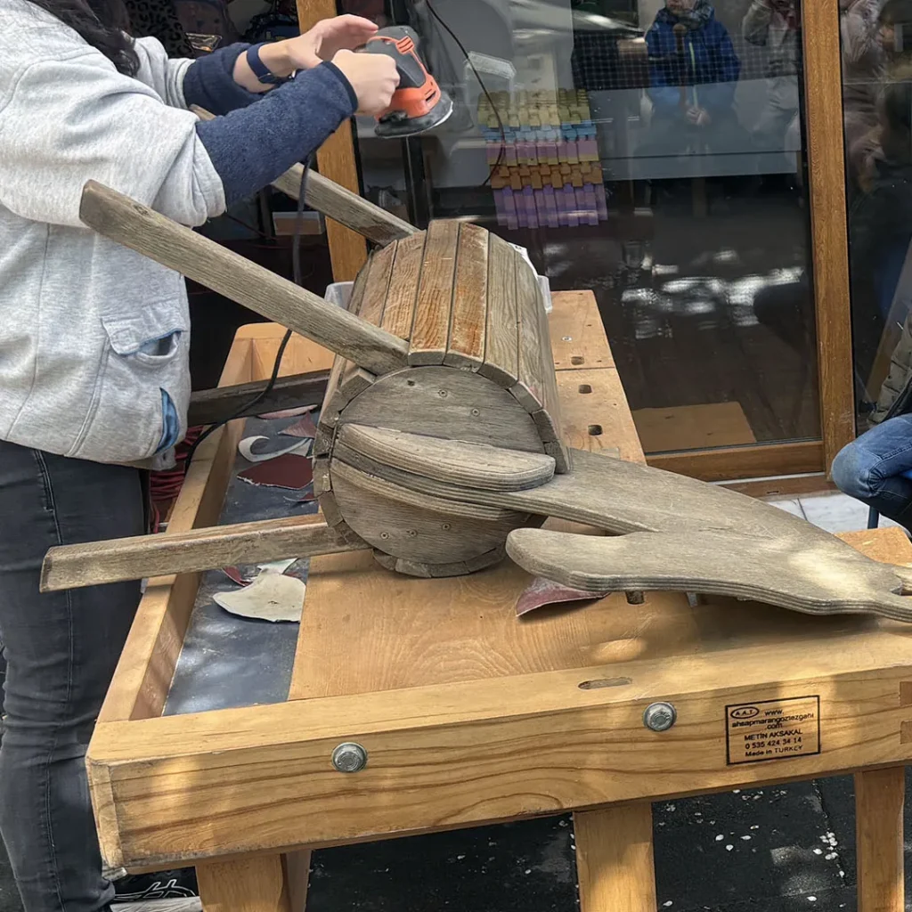 Eine Begleiterin schleift das Holzpferd mit einer elektrischen Handschleifmaschine ab